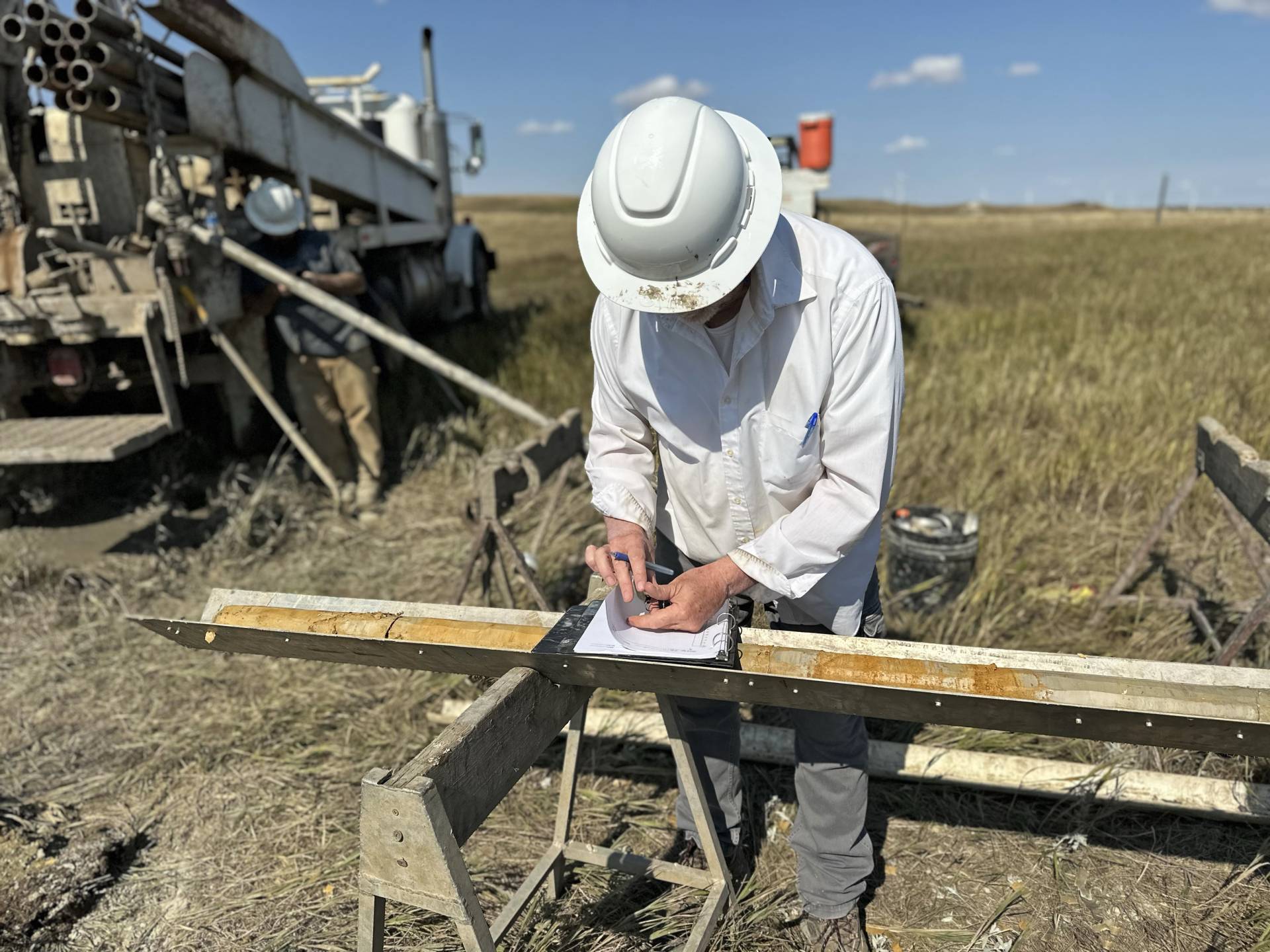 A man wearing a hard hat is writing in a notebook next to a truck in a field. 